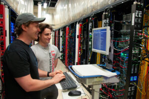 Two people work at a computer surrounded by servers in a data center.